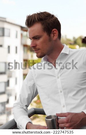Attractive young man standing on a balcony holding a coffee mug. Wearing a white shirt.