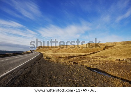 Winding Rural Road with No Cars Single lane, paved road winding through rural hills on a sunny day with bright blue skies and smooth clouds. The road is empty. Setting: Icelandic Ring Road.