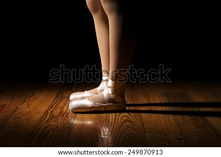 Closeup of a ballerina\'s feet in pointe shoes on a vintage, wood floor with black background. Feet are resting on ground.