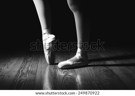 Closeup of a ballerina\'s feet in pointe shoes on a vintage, wood floor with black background. Black and white image with drama. One foot is pointed and the other is flat.