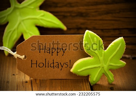 Brown Organic Label With English Text Happy Holidays On Wooden Background With Two Leaf Cookies And Frame