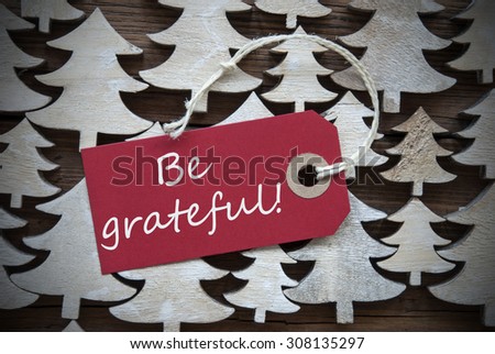 Red Christmas Label With Ribbon On Wooden Christmas Trees Background. Vintage Or Rustic Style. Label With English Text Be Grateful For Christmas Or Season Greetings.Close Up Or Macro