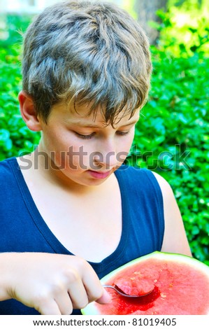 The boy is eating in the courtyard of a red, sweet watermelon with a spoon