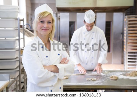 Baker posing in bakery or bakehouse. Another baker is working in the background