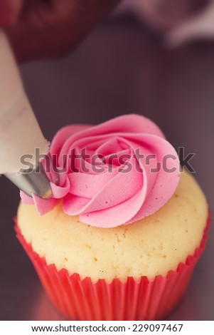 Close up pink rose frosted cupcake being iced with piping bag