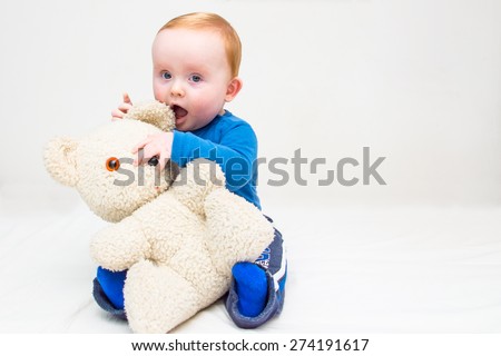 Seven month old white baby boy, with blue eyes and red hair, sitting on a white mattress and plays with his teddy bear. He is wearing a blue bodysuit and blue socks. Isolated on white.