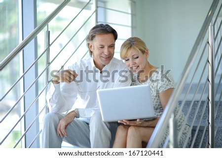 Confident business partners sitting on stairs in office building and discussing work, computer, smiling, mature businessman