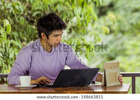 young man in purple t-shirt  keying notebook holding tablet with coffee cup at terrace