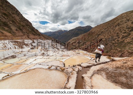 Worker carrying a big sack of salt, on the terraced salt pans in Maras, Urubamba Valley, Peru. Concept of manual work in developing countries. Wide angle image, rear view, unrecognizable person.
