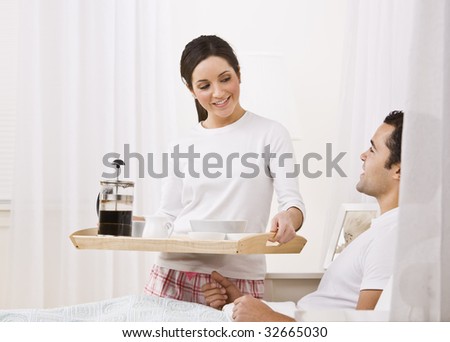 A beautiful young woman serving a breakfast tray to a man lying in bed.  They are smiling happily at one another. Horizontally framed shot.