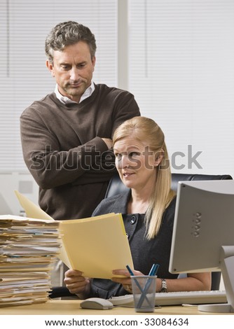 A businessman is standing in an office behind the receptionist, and is looking over her shoulder to the file in her hands.  She is looking away.  Vertically framed shot.
