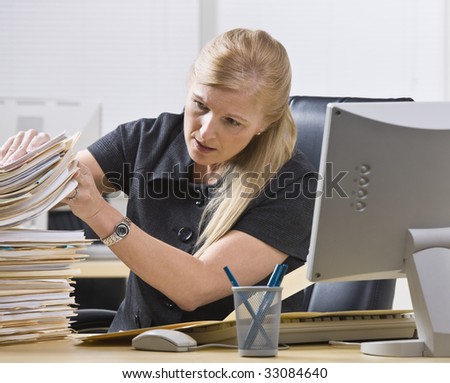 A businesswoman is seated at a desk in an office and is looking through a stack of paperwork.  Horizontally framed shot.