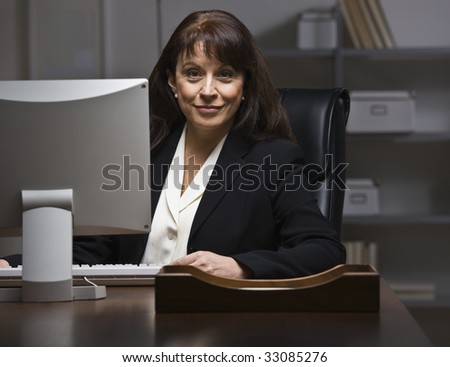 Attractive businesswoman sitting at desk in suit, behind monitor. Looking at camera. Horizontal