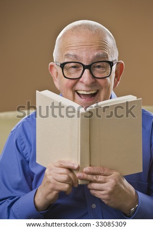 Laughing senior male reading a book on a couch. Vertical