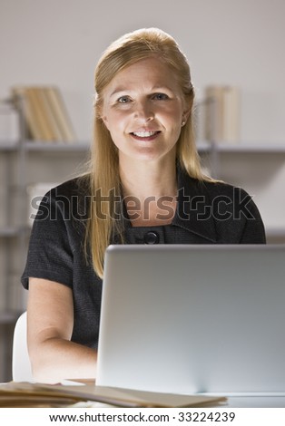 A businesswoman is sitting at a desk in an office.  She is working on a computer and smiling at the camera.  Vertically framed shot.