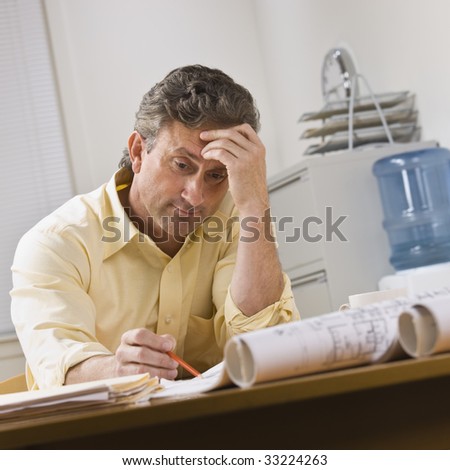 A businessman is working on blueprints in an office.  He is looking away from the camera.  Square framed shot.
