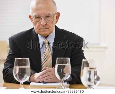 An elderly businessman is seated at a desk in an office and is looking at the camera.  Horizontally framed shot.