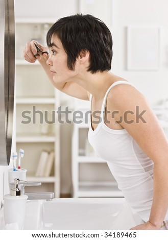 A woman is standing in front of the mirror and trimming her bangs.  Vertically framed shot.