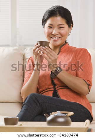 A woman is seated on her sofa, and drinking tea out of a tea cup.  She is smiling at the camera.  Vertically framed shot.