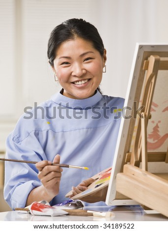 A woman is painting a picture on an easel.  She is smiling at the camera.  Vertically framed shot.