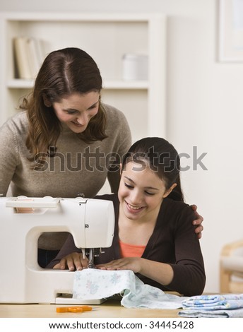 A beautiful teenage girl sewing on a sewing machine while her mother watches over her shoulder.  They are smiling.  Vertically framed shot.