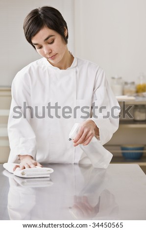 A chef is cleaning a counter in a professional kitchen with a bottle of solution and a rag.  Vertically framed shot.
