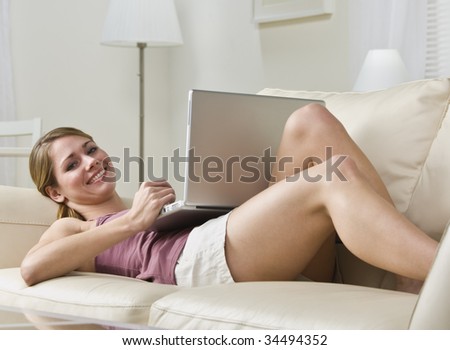 Attractive blonde woman lying on couch with laptop, smiling at camera. Horizontal