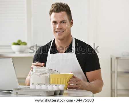 A young man with flour on his face is mixing ingredients in his kitchen.  He is smiling at the camera.  Horizontally framed shot.