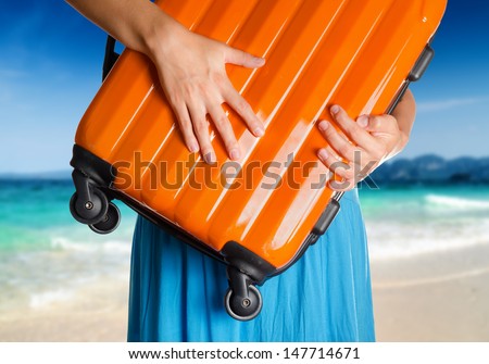 Woman in blue dress holds orange suitcase in hands on the beach background.