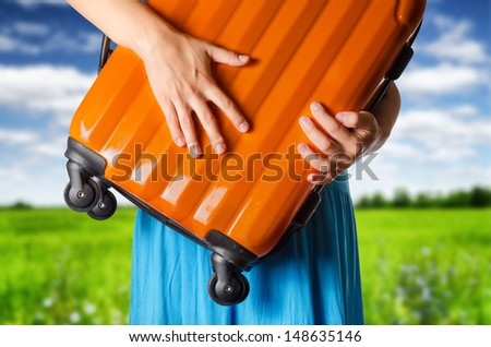 Woman in blue dress holds orange suitcase in hands on the field background.