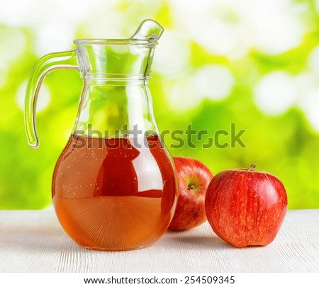 Jug of apple juice and red apples on nature background. Half full pitcher. Eco food rich in minerals and vitamins. Product of organic farming.