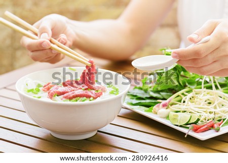 Young woman eating and enjoying the Pho Bo in street cafe of Vietnam. The Pho Bo is a traditional Vietnamese beef noodle soup with garnish of leaves of cilantro and Asian basil. Popular street food.