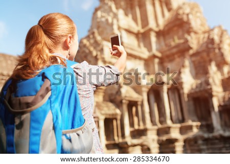 Closeup view of young female tourist with smartphone taking picture of upper gallery at ancient temple complex Angkor Wat in morning sun. Siem Reap, Cambodia.