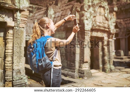 Young female tourist with blue backpack and smartphone taking picture of mysterious ruins in the ancient Preah Khan temple in Angkor. Siem Reap, Cambodia. Toned image.