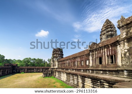 Wall of gallery and one of towers of main Temple Mountain of ancient temple complex Angkor Wat in Siem Reap, Cambodia. Blue sky and woods in background. Angkor Wat is a popular tourist attraction.