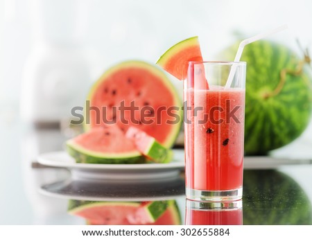 Glass of fresh red watermelon juice on table in kitchen. Ripe watermelons and slices in background. Healthy eco sweet food rich in vitamins. Popular product of organic farming.