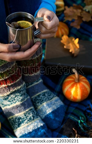 Female hands with hot tea cup and female legs in warm knitted socks on plaid background, book, autumn leaves and small pumpkin