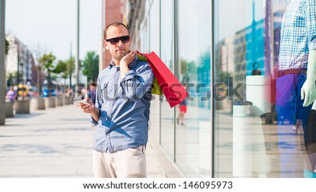 Young man shopping in the mall with many colored shopping bags in his hand. He is holding a phone. In the background window dressing.
