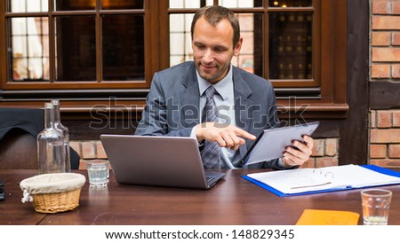 Hard working smiling businessman in restaurant with laptop and pad.