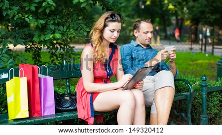 Young couple sitting on a bench with colorful shopping bags and mobile phone and tablet.