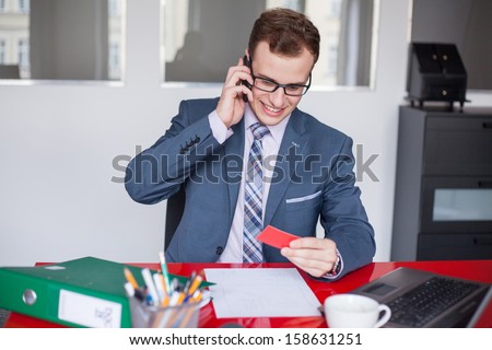 Young businessman holding mobile phone and red business card. He is sitting in his office.