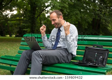 Excited businessman sitting on a bench in park with laptop.