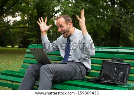 Excited businessman sitting on a bench in park with laptop.
