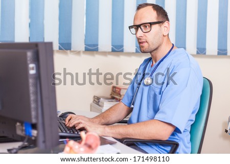 Doctor working on his computer and with mobile phone in the office. He is wearing blue uniform (surgeon uniform)