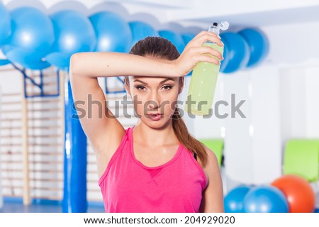 Young girl drinking isotonic drink, gym. She is wiping sweat.
