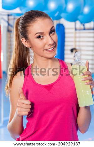 Young girl drinking isotonic drink, gym. She is happy and full of positive emotion. Thumb up