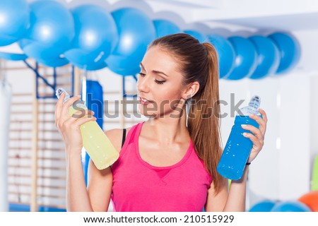 Young girl with two isotonic drink (yellow, blue), gym. She is considering which one to choose
