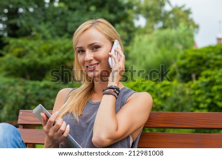 Sensual blonde woman sitting in park on wooden bench. She is using mobile phone and tablet pc. Outdoor photo. She looks relaxed
