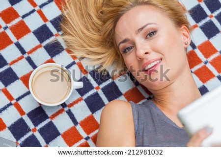 Sensual blonde woman lying in park on blanket. She is using white tablet pc. Outdoor photo. She looks relaxed