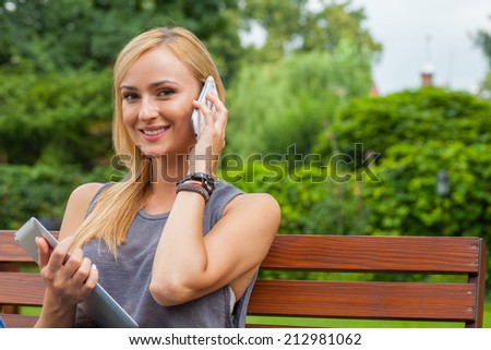 Sensual blonde woman sitting in park on wooden bench. She is using mobile phone and tablet pc. Outdoor photo. She looks relaxed
